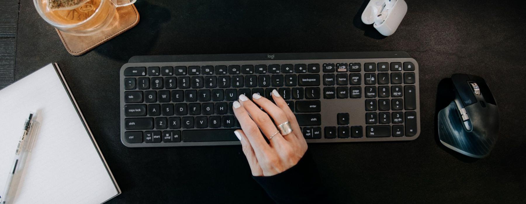 woman's hand on a keyboard surrounded by office items and a cup of tea on a coaster